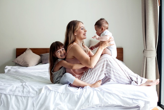 Young mom with her 5 years old daughter and 4 months old baby dressed in pajamas are relaxing and playing in the bed at the weekend together, lazy morning